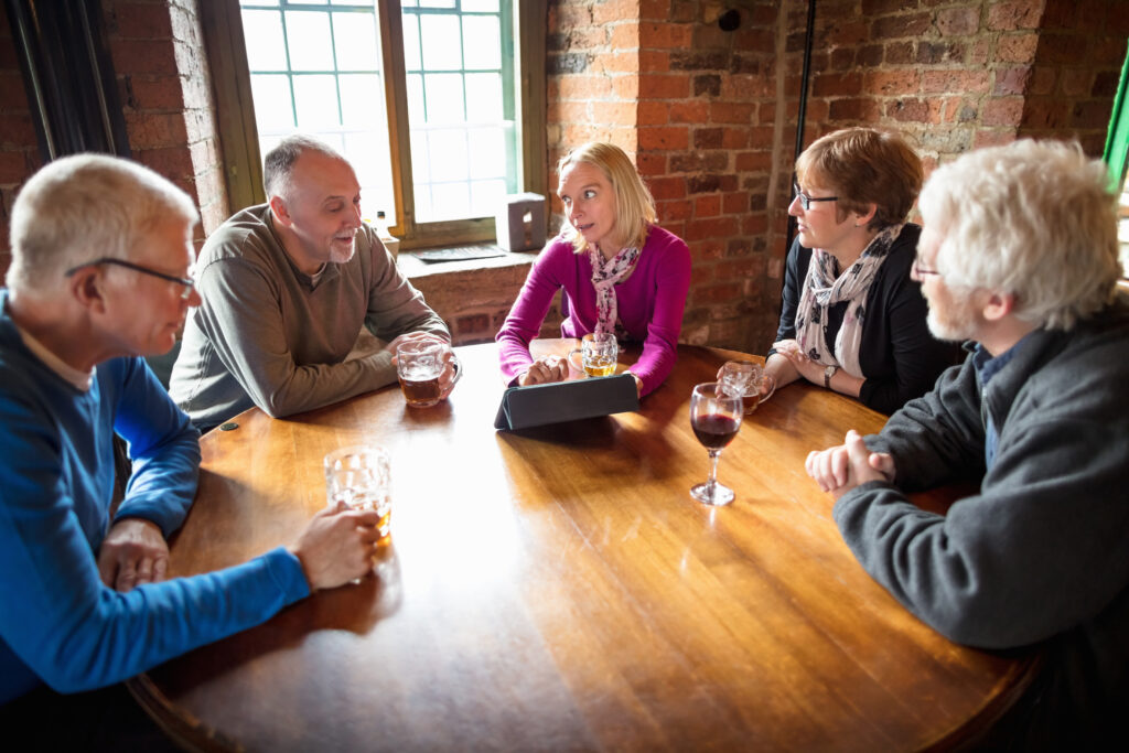 High angle view of a group of men and women in their 50s and 60s sitting around a large table in a pub, drinking wine and beer. They are looking at the screen of a tablet computer.